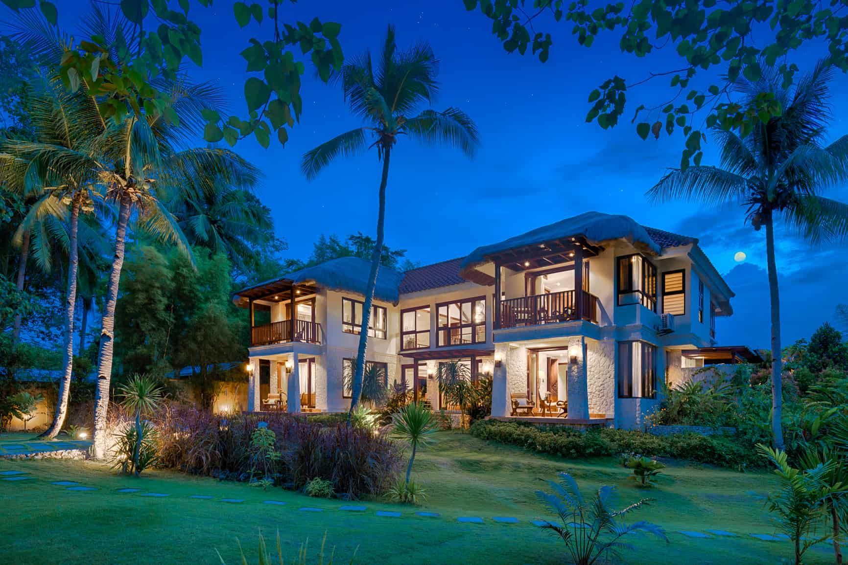 Luxury beachfront villa at dusk, illuminated, with palm trees and a grassy lawn.