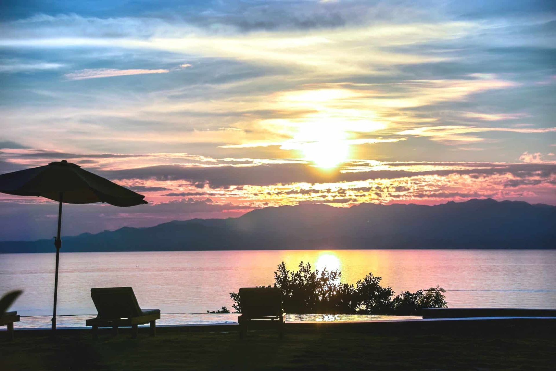 Sunset over calm water with lounge chairs and umbrella silhouetted on a beach.