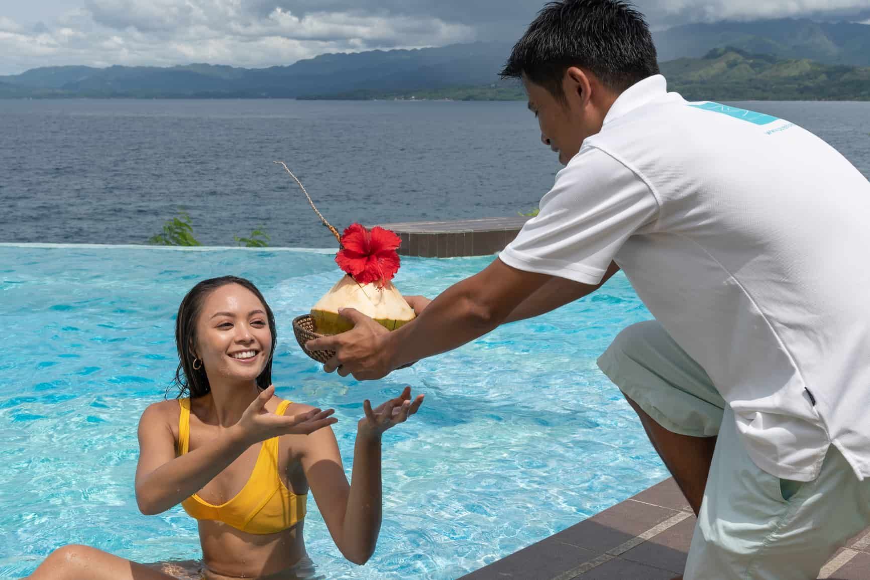 Person in pool receives a coconut drink from a person on the pool deck. Ocean in background.