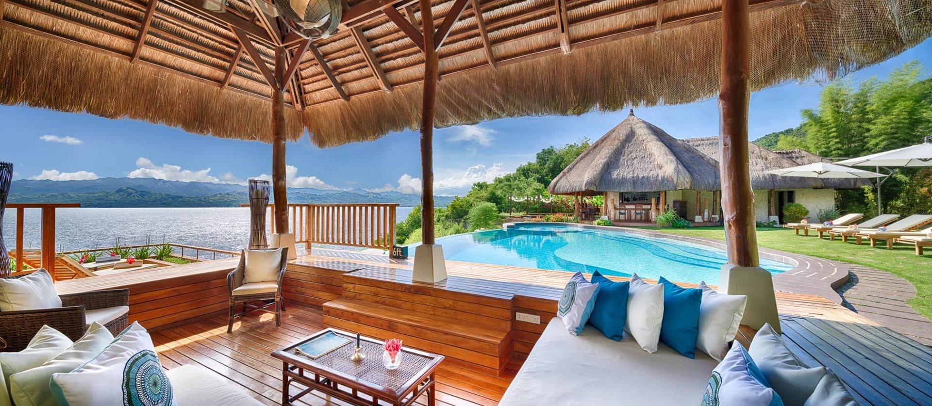 Lush resort view with thatched roof, pool, lake, and seating with cushions.