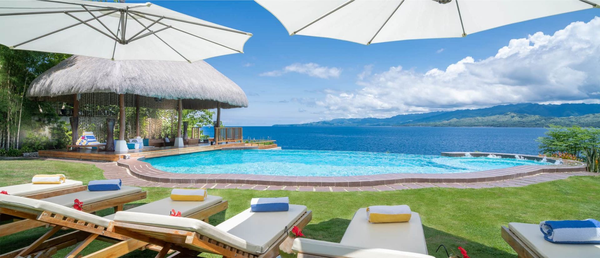 Lounge chairs by an infinity pool overlooking a body of water under a sunny sky.