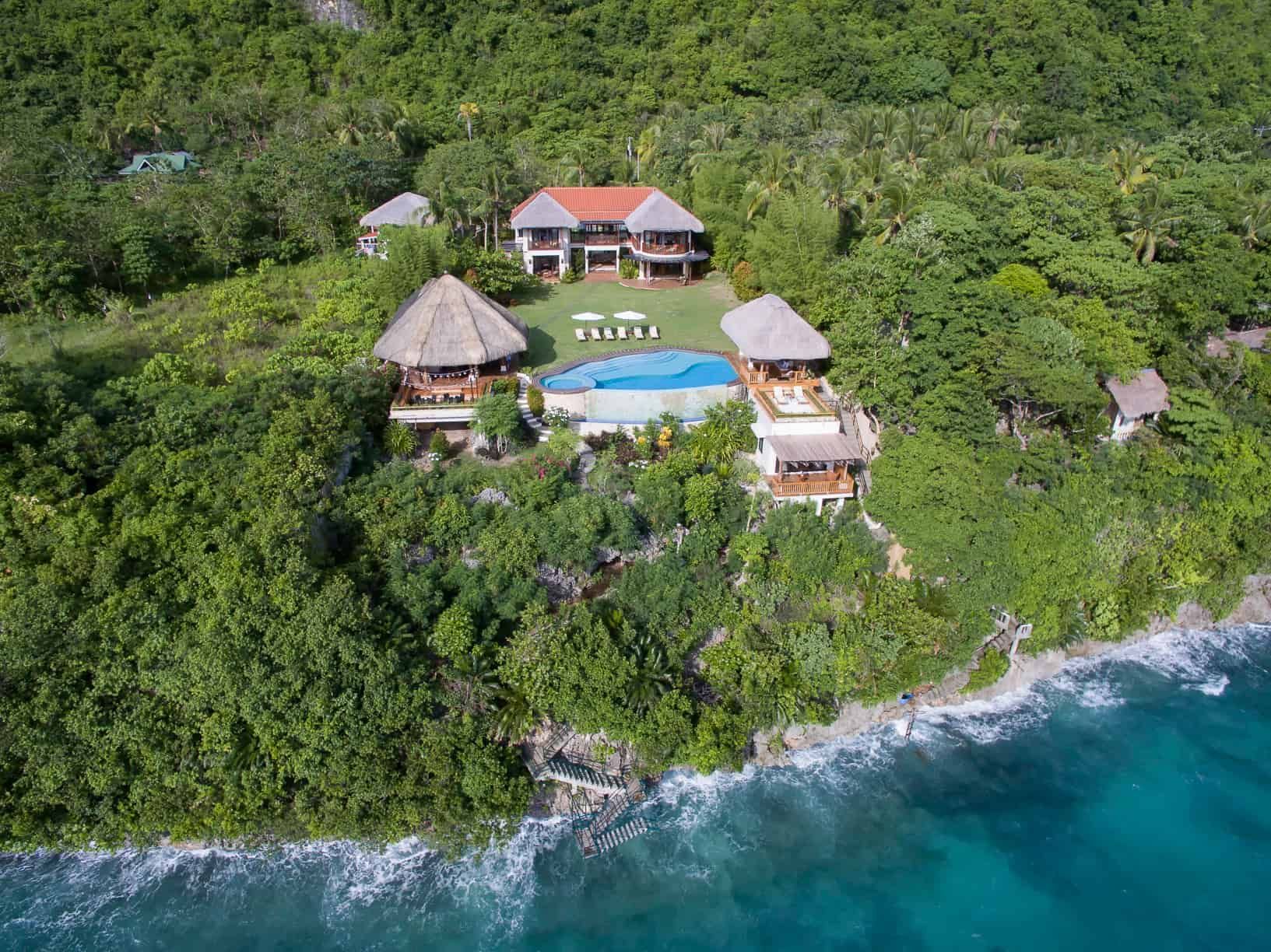 Aerial view of a coastal resort with buildings, a pool, and ocean, surrounded by lush green vegetation.
