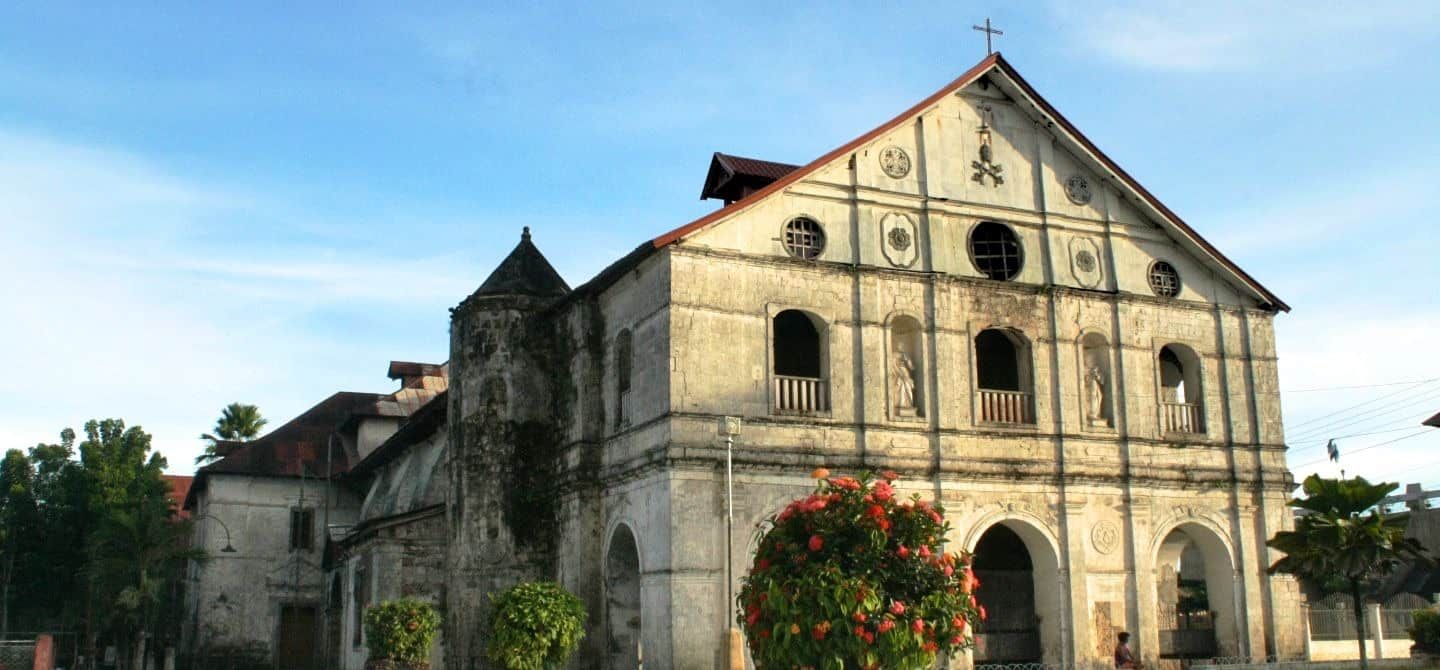 Exterior view of an aged, stone church with a cross on top, blue sky backdrop.