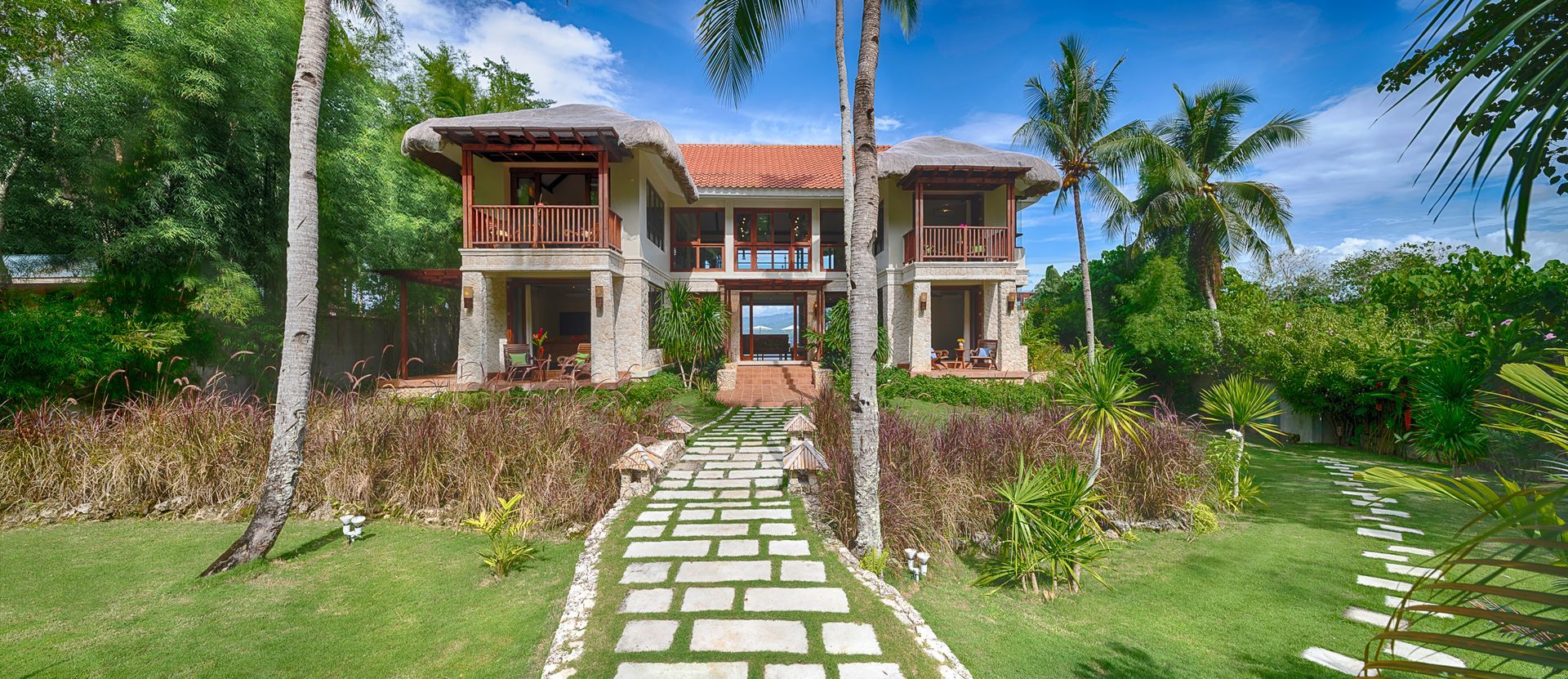Two-story tropical home with balconies, a stone path, and lush greenery under a blue sky.
