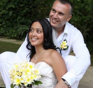 Bride and groom in white attire, smiling, with a bouquet.