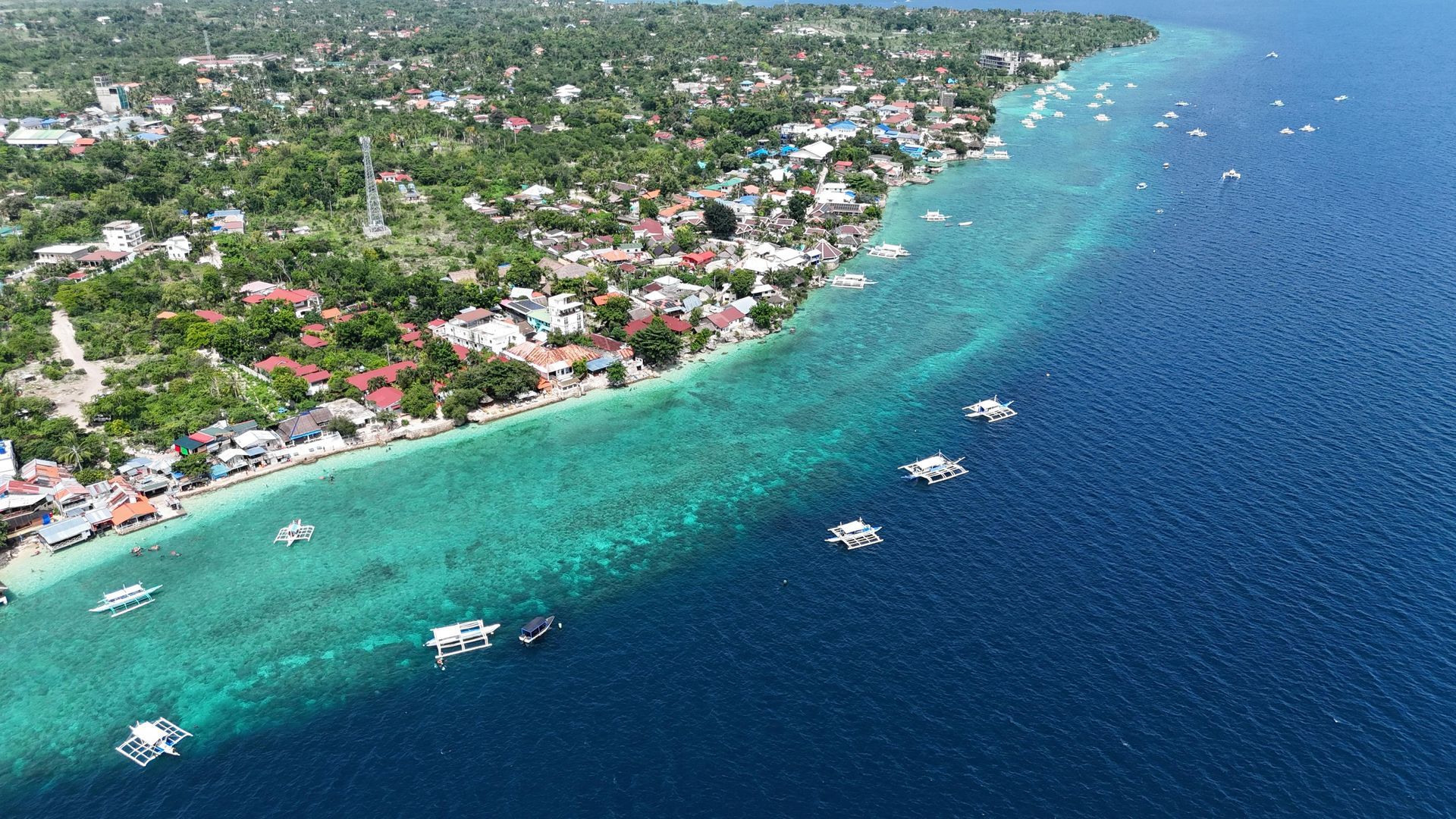 Aerial view of a coastal town with clear turquoise water and boats; lush green vegetation inland.