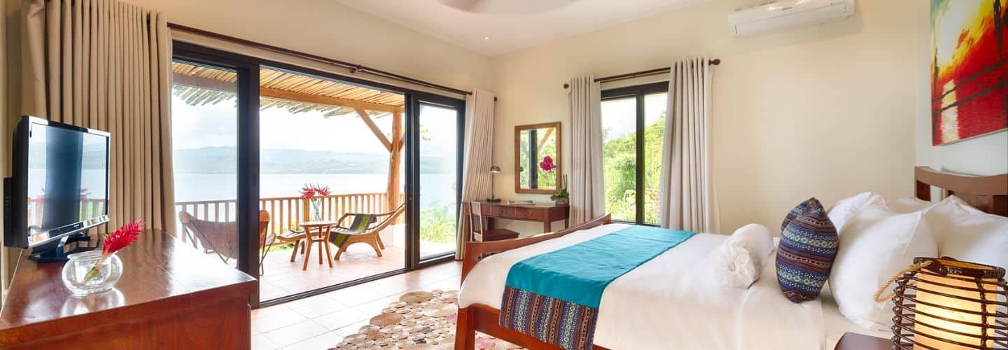 Bedroom with balcony overlooking a body of water; white bed with teal blanket; wooden furniture.