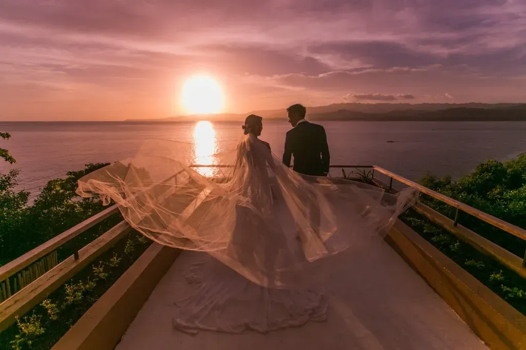 Bride and groom watching sunset over ocean. Train of wedding dress blowing in the wind.