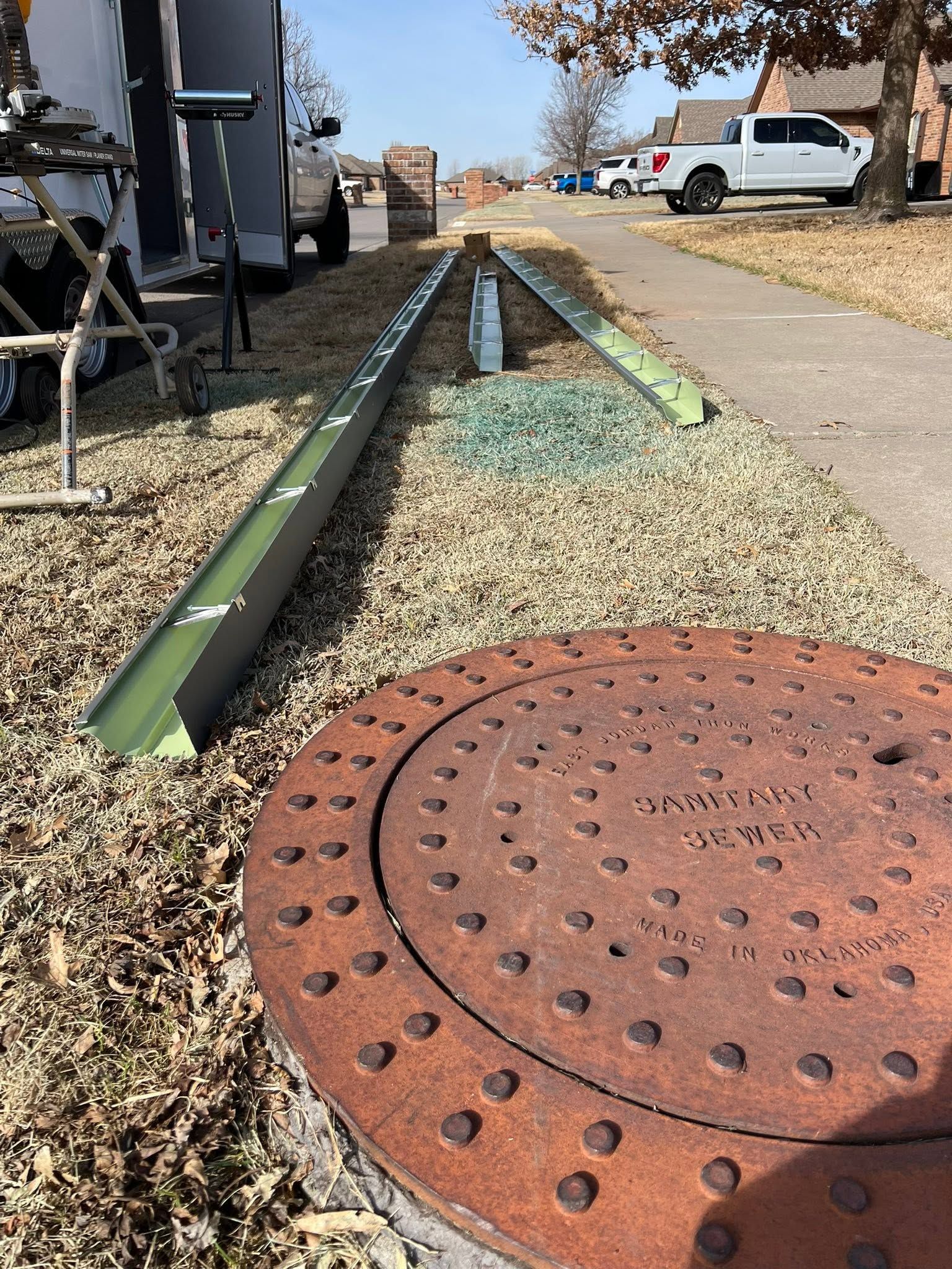 Rusty manhole cover beside a narrow concrete drainage channel in a dry roadside lot, with a white truck in the background