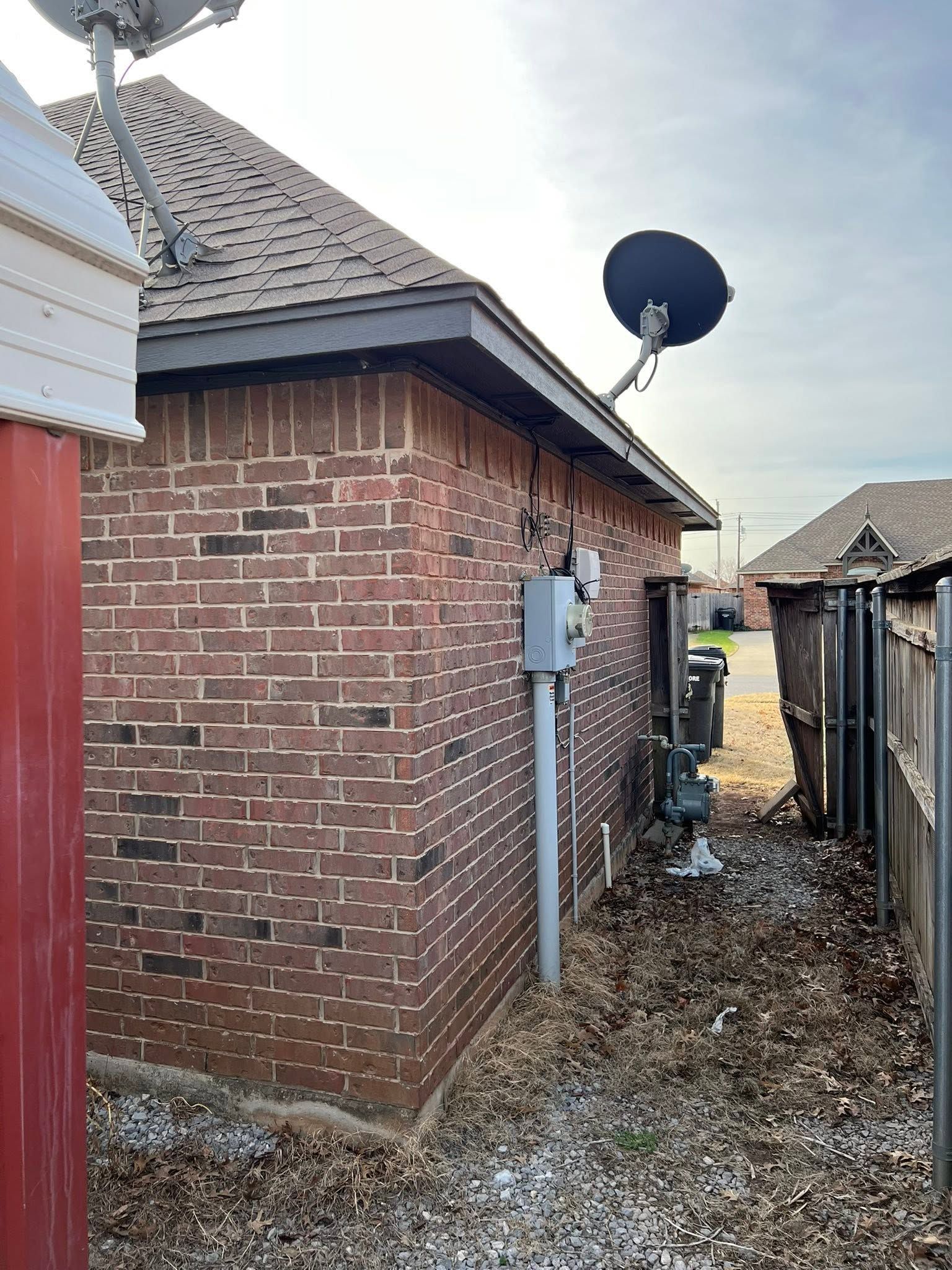 Brick house side yard with satellite dishes, utility box, and a narrow gravel path by a wooden fence