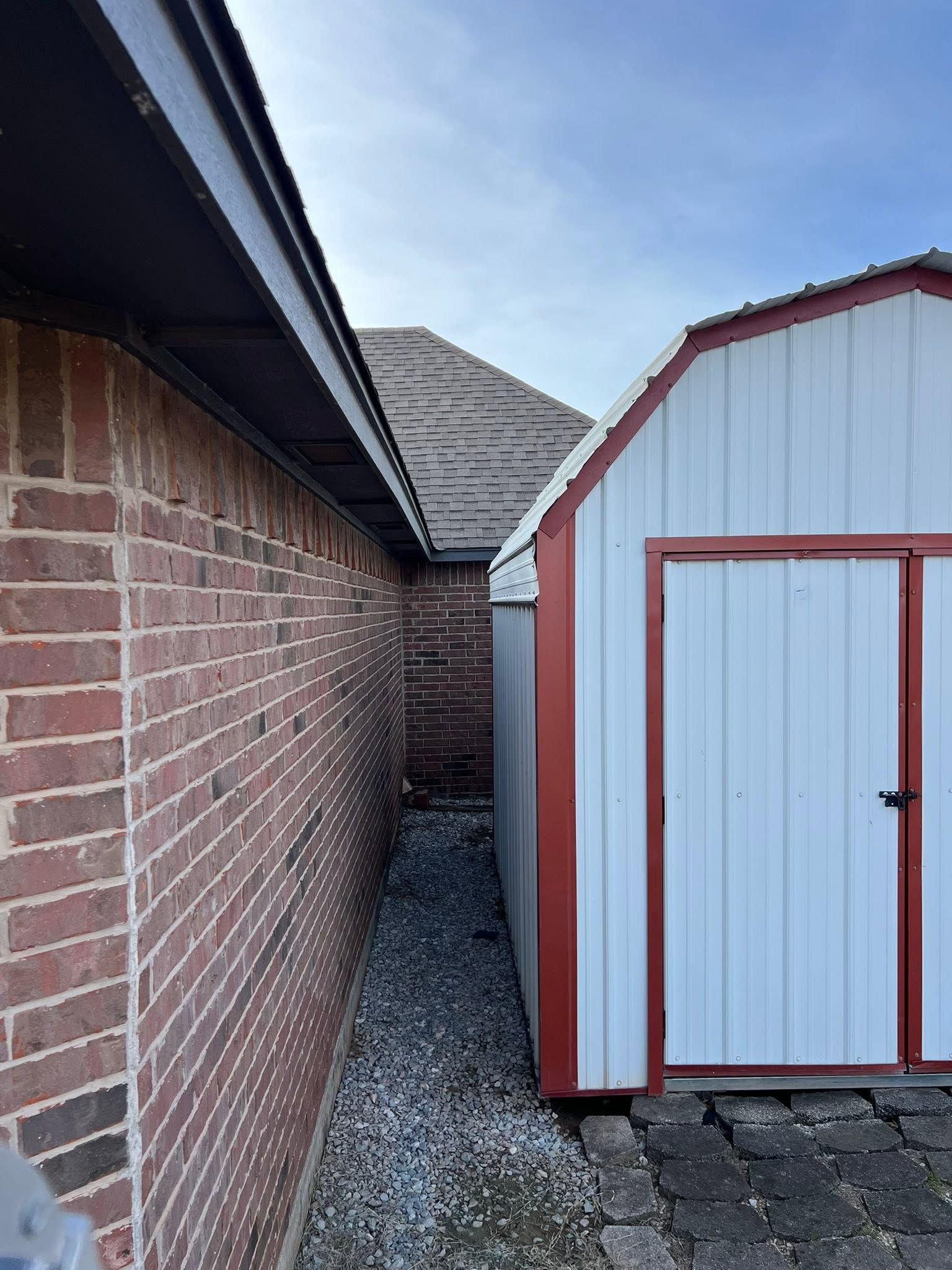 Narrow gravel alley between a brick wall and a white shed with red trim under a cloudy sky