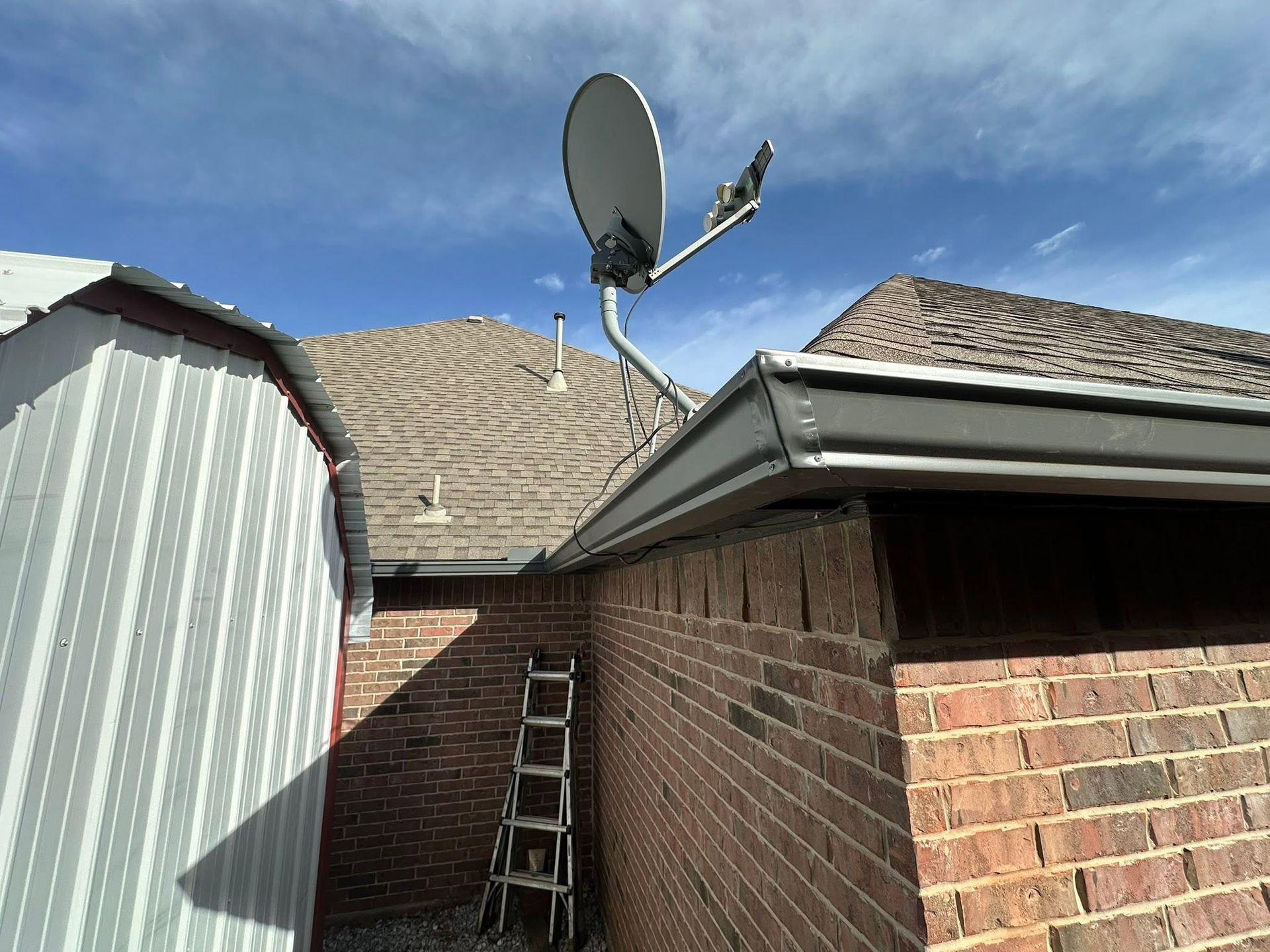 Brick house roof with satellite dish and ladder beside a shed under a blue sky