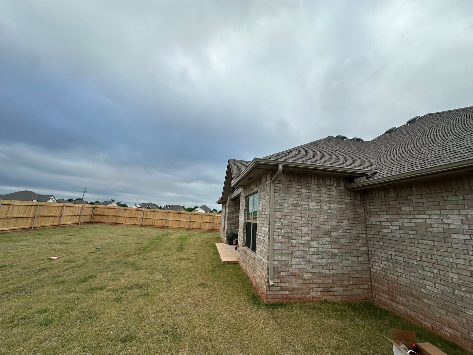 Backyard with grass, wooden fence, and a brick house under a cloudy sky
