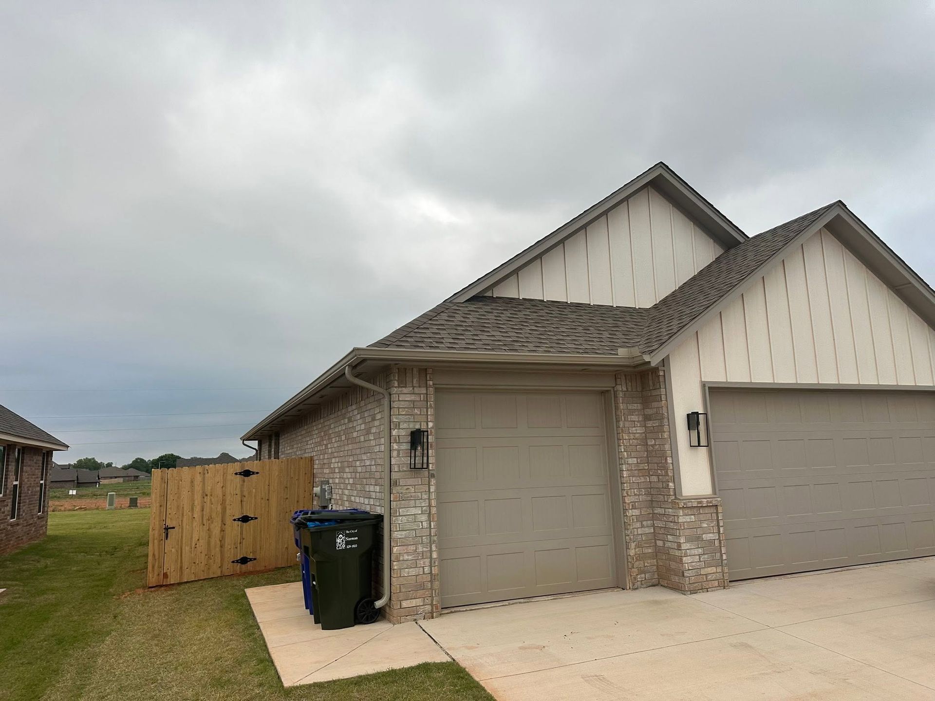 Suburban duplex with two garage doors, beige siding, and a trash bin by the driveway under cloudy skies