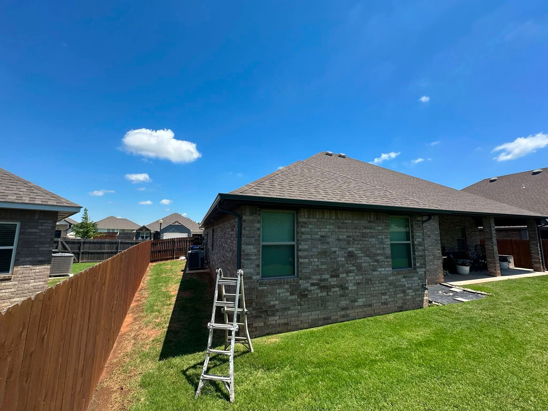 Suburban backyard with stone houses, green lawn, wooden fence, and ladder under a blue sky