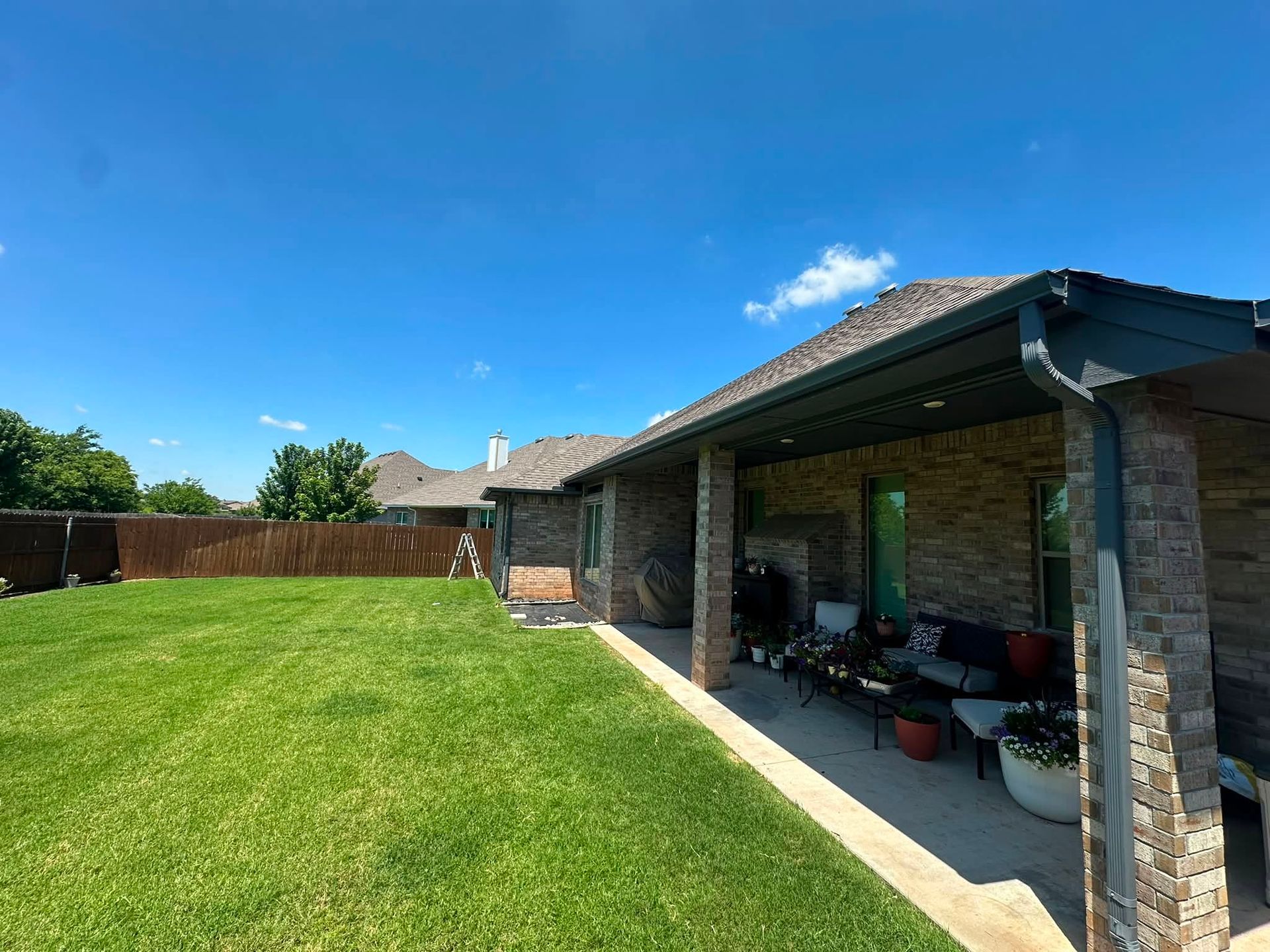 Backyard with green lawn, brick patio, and covered outdoor seating under a blue sky