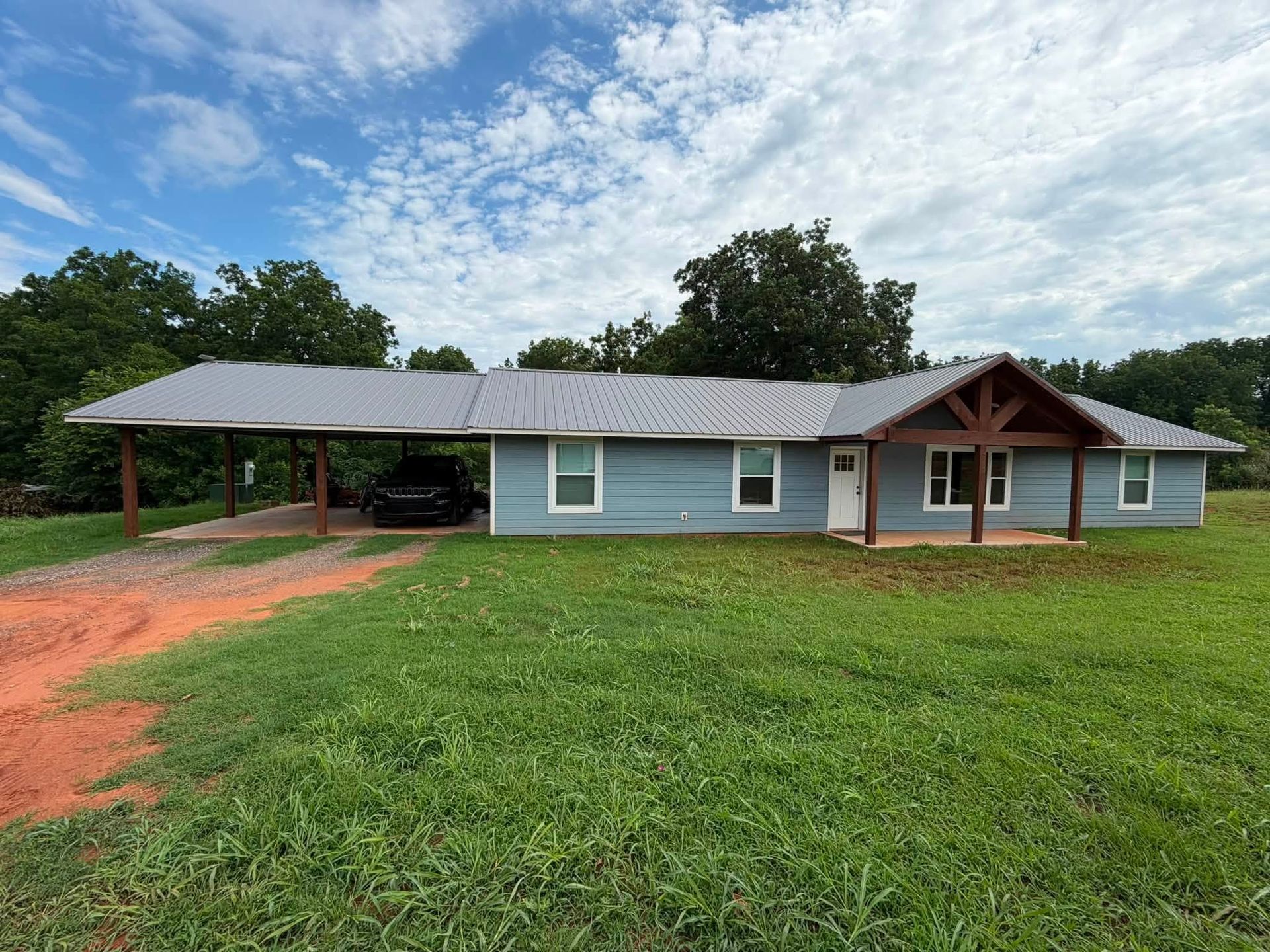 Single-story blue house with gray roof, red porch, and carport on a grassy lot under a cloudy sky