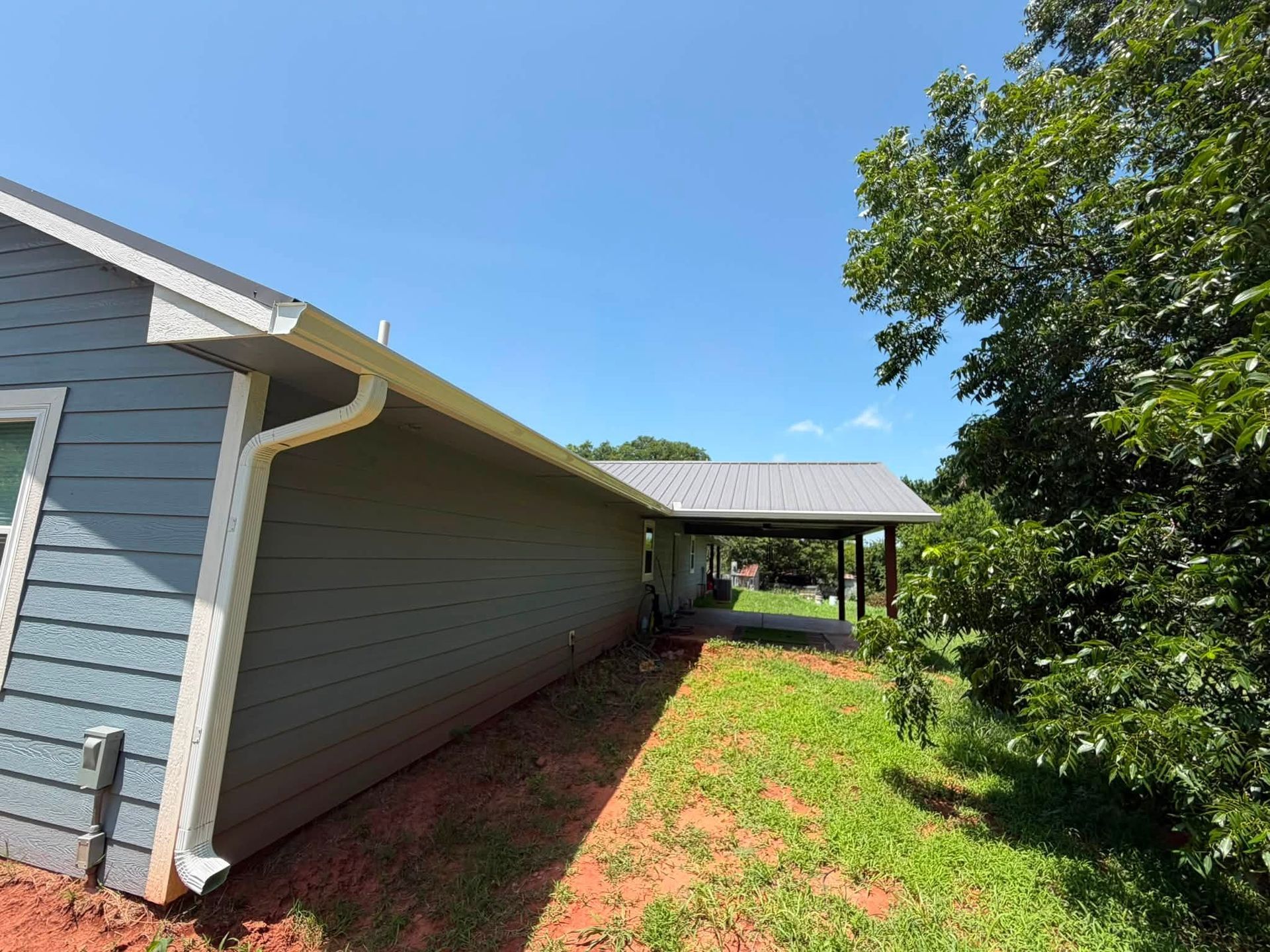 Side yard beside a gray house with a covered patio, grass, and trees under a blue sky