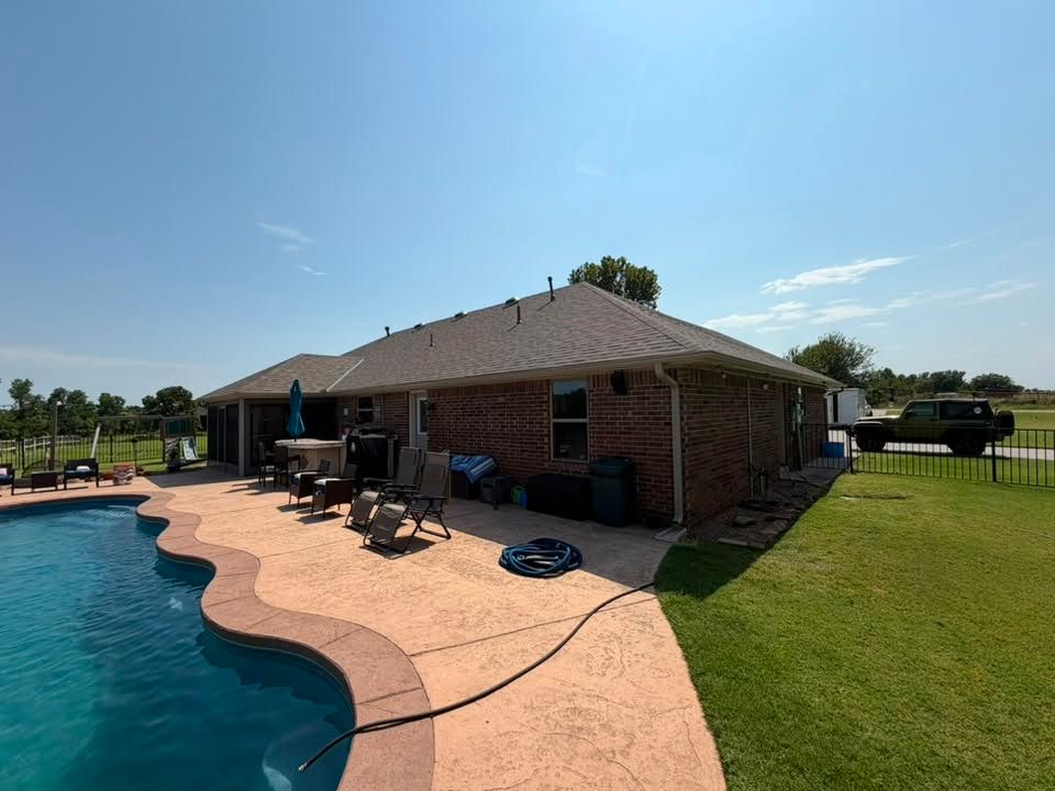 Backyard pool with brick house, patio chairs, green lawn, and sunny blue sky