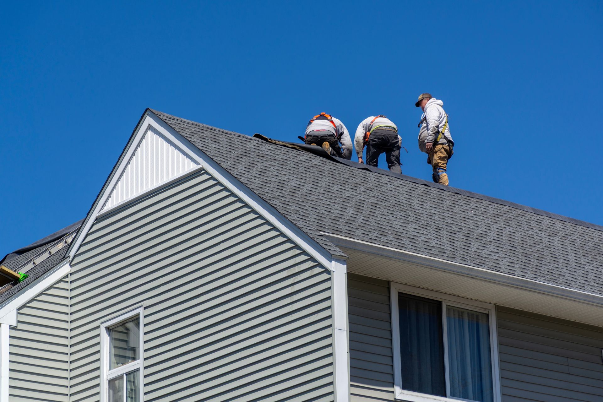 Roofers repairing gray shingle roof on a white house under a blue sky