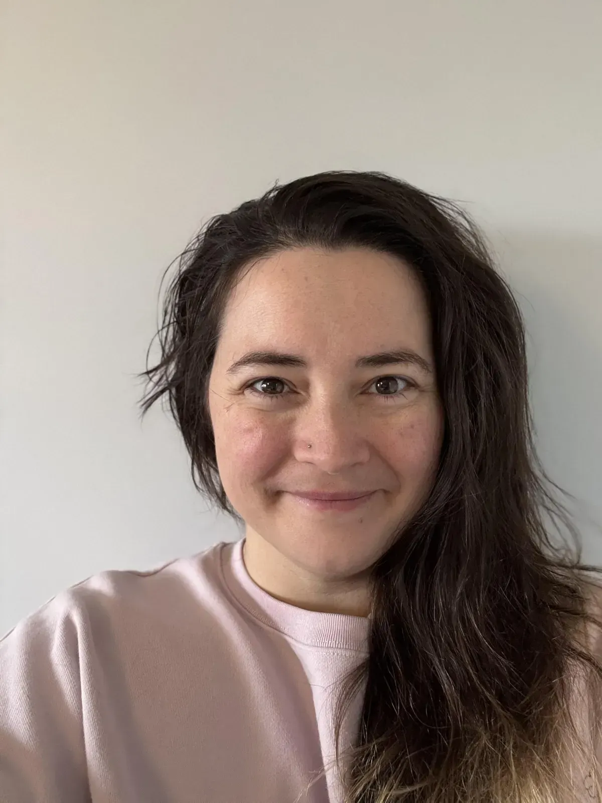 Woman with brown hair and pink sweater smiles at the camera against a plain white wall.