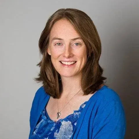 Woman with brown hair smiling, wearing a blue cardigan and pendant necklace, against a gray backdrop.