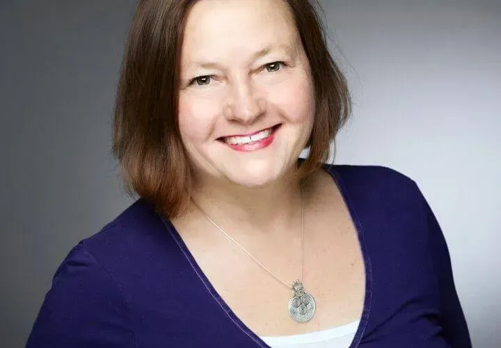 Woman with brown hair, smiling, wearing a blue top and silver necklace.