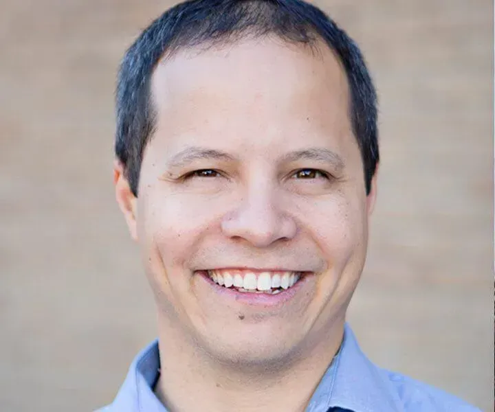 Man with dark hair smiling, wearing a blue shirt, against a light brown background.