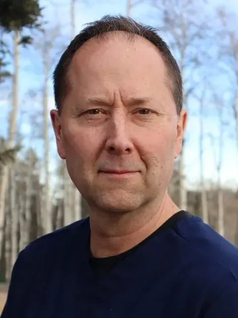 Man with short brown hair, blue shirt, neutral expression, set against a blurred outdoor background of trees.