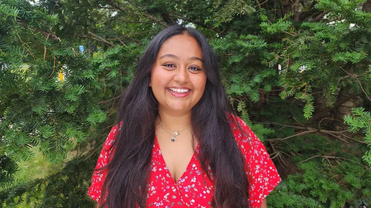 Woman with long dark hair, wearing a red floral top, smiles broadly in front of green foliage.