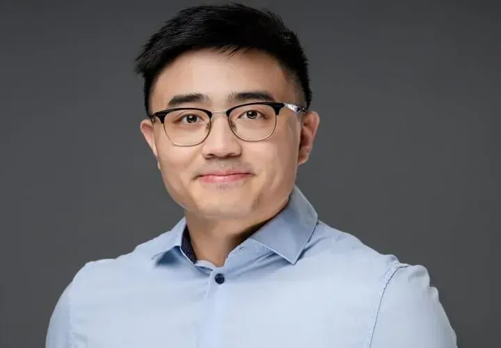 Man with glasses, light blue shirt, smiling, posing in front of a gray backdrop.