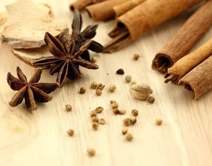 Close-up of spices: star anise, cinnamon sticks, coriander seeds, and cardamom pod on a light wood surface.