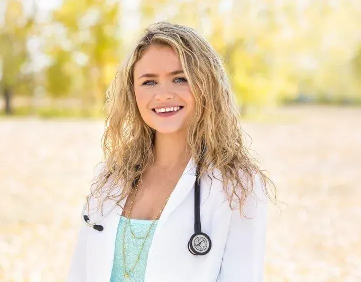 Woman with blonde curly hair smiles wearing a white coat and stethoscope outdoors with blurred foliage.