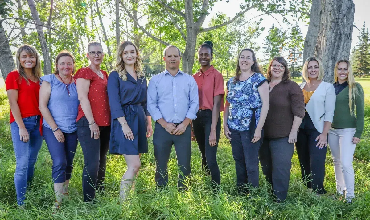 Group of people standing outdoors, smiling. They wear various colored tops and stand in front of a tree.