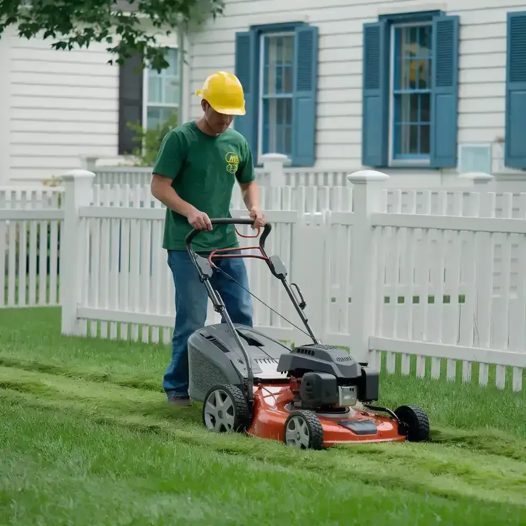 Worker mowing grass with a red lawn mower beside a white fence and blue-shuttered house