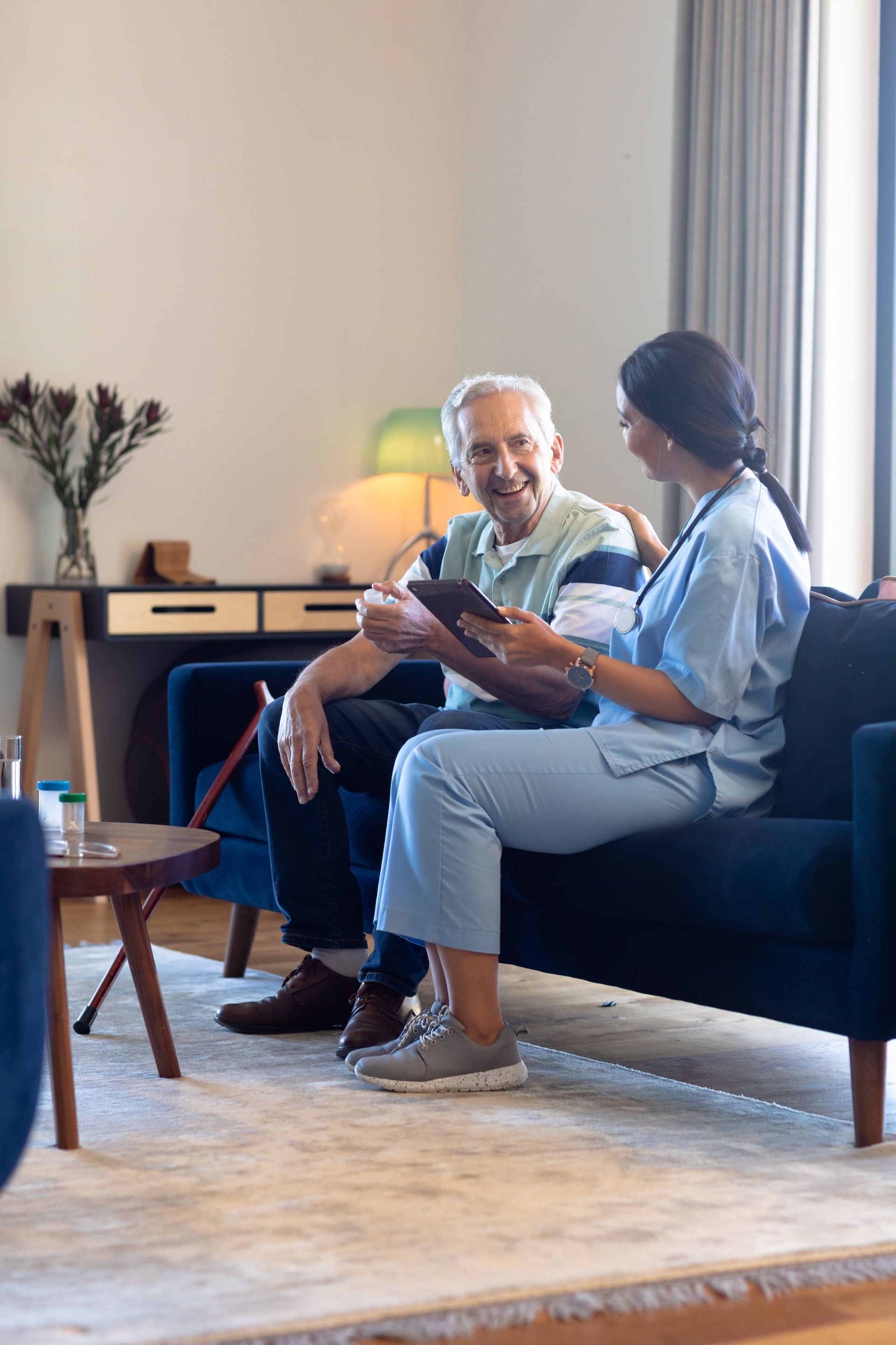 Caregiver in blue scrubs showing a tablet to an older person seated on a navy couch in a living room.