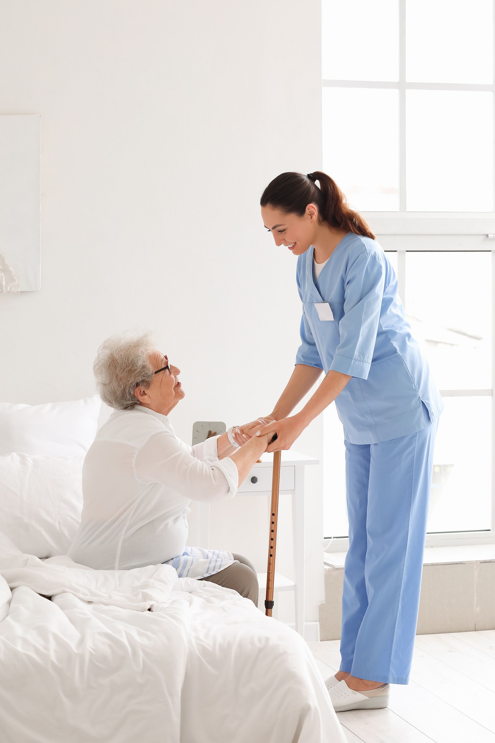 Nurse helps elderly person sit up in bed, smiling in a bright room.