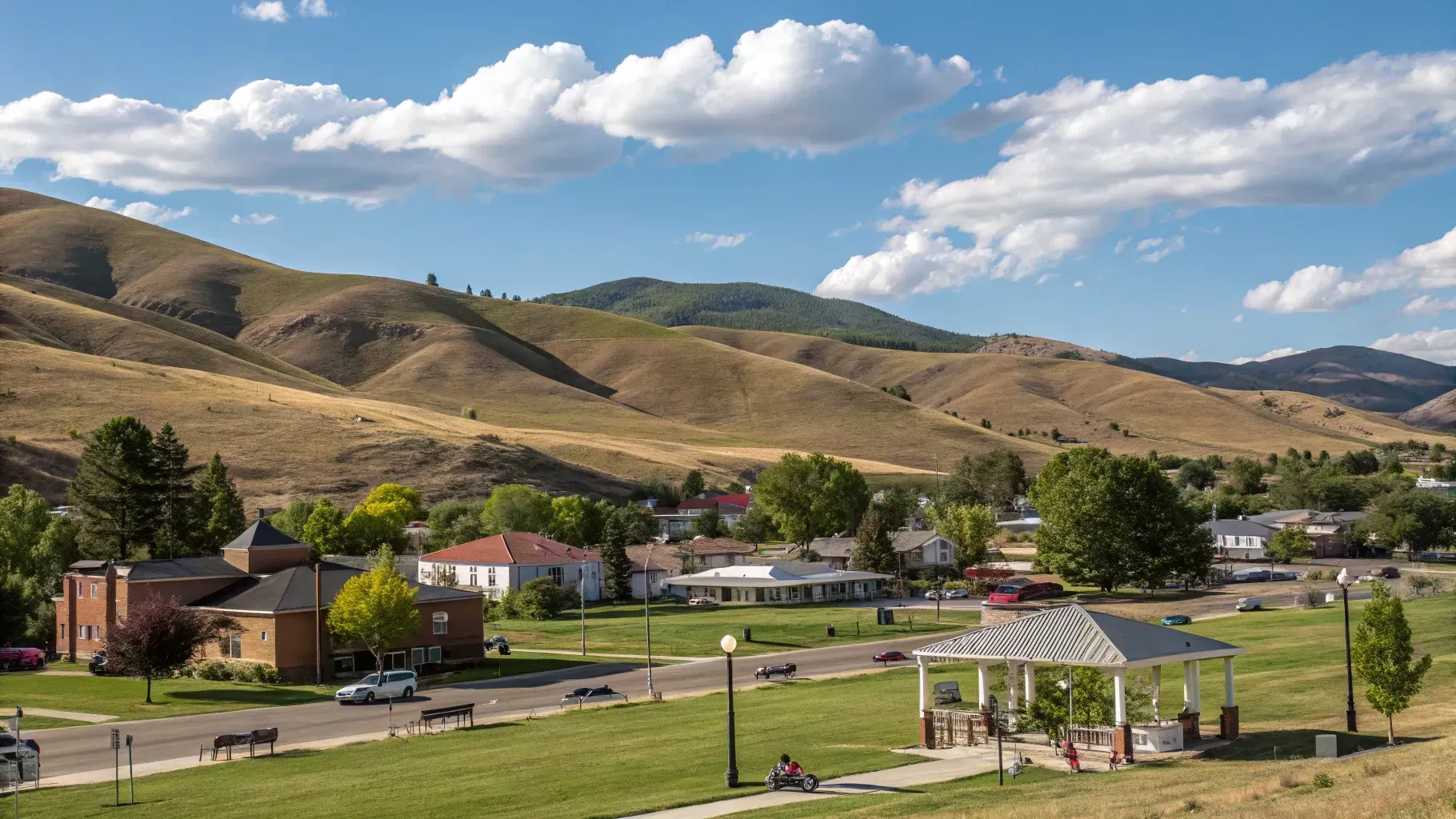 Town nestled in valley with green lawn and gazebo; rolling brown hills in background.