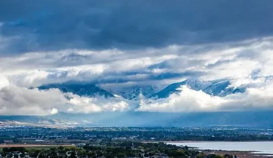 Mountains peeking through storm clouds over a town and lake. Blue sky, white clouds, green landscape.