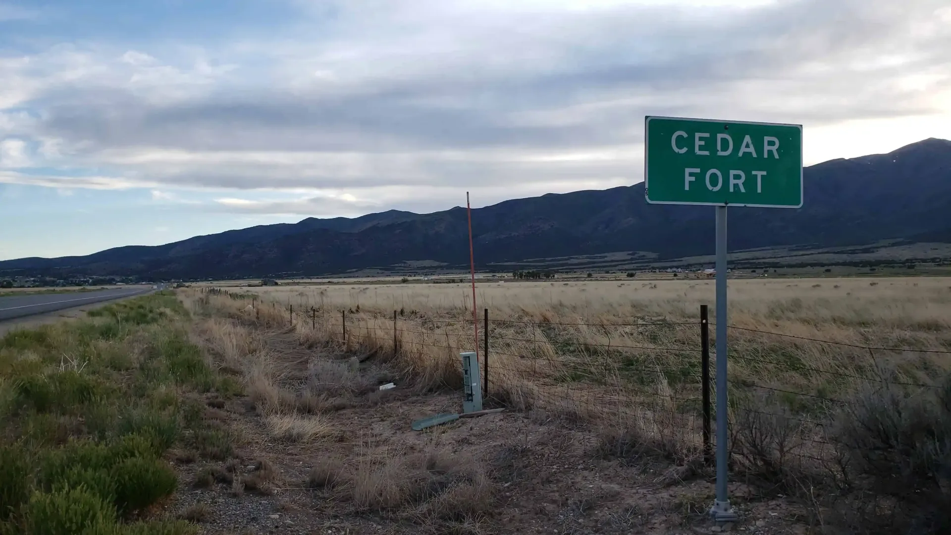 Green sign for Cedar Fort next to a road with mountains in the background.