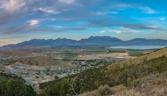 Mountain range overlooks a valley with town and fields, under a cloudy blue sky.