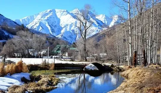 Snowy mountains reflected in a clear river, with trees and small buildings in the valley.