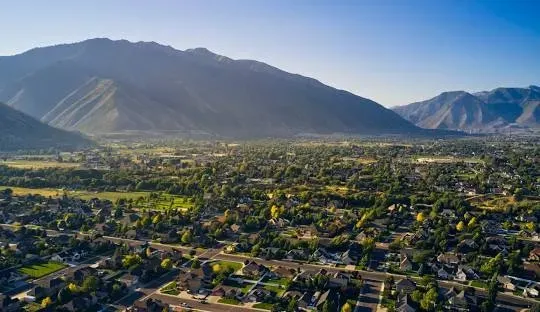 Aerial view of a valley town with houses and trees, mountains in the background under a blue sky.