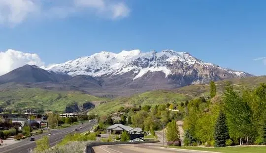 Snow-capped mountain overlooking a lush green valley with a road and scattered buildings under a blue sky.