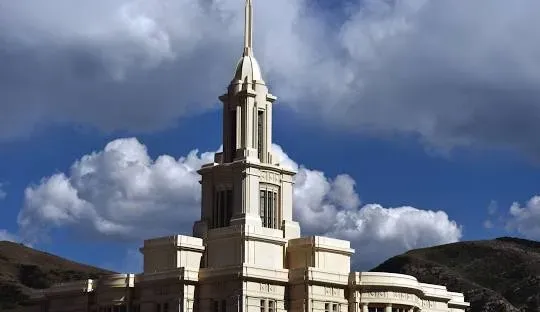 White temple with steeple against a blue sky with puffy clouds, set on a mountainside.