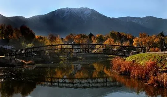 A bridge arches over a lake, reflecting the structure and fall foliage with a mountain backdrop.