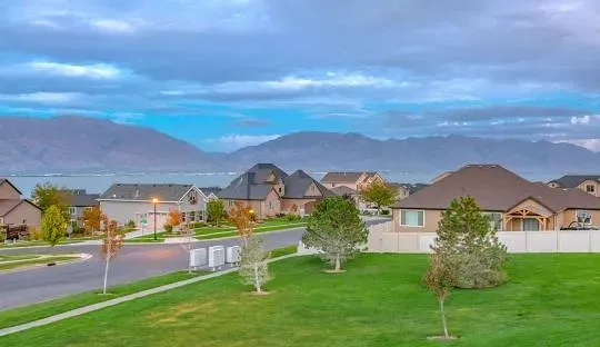 Suburban neighborhood with houses, trees, green lawns, road, and distant mountains under a cloudy sky.