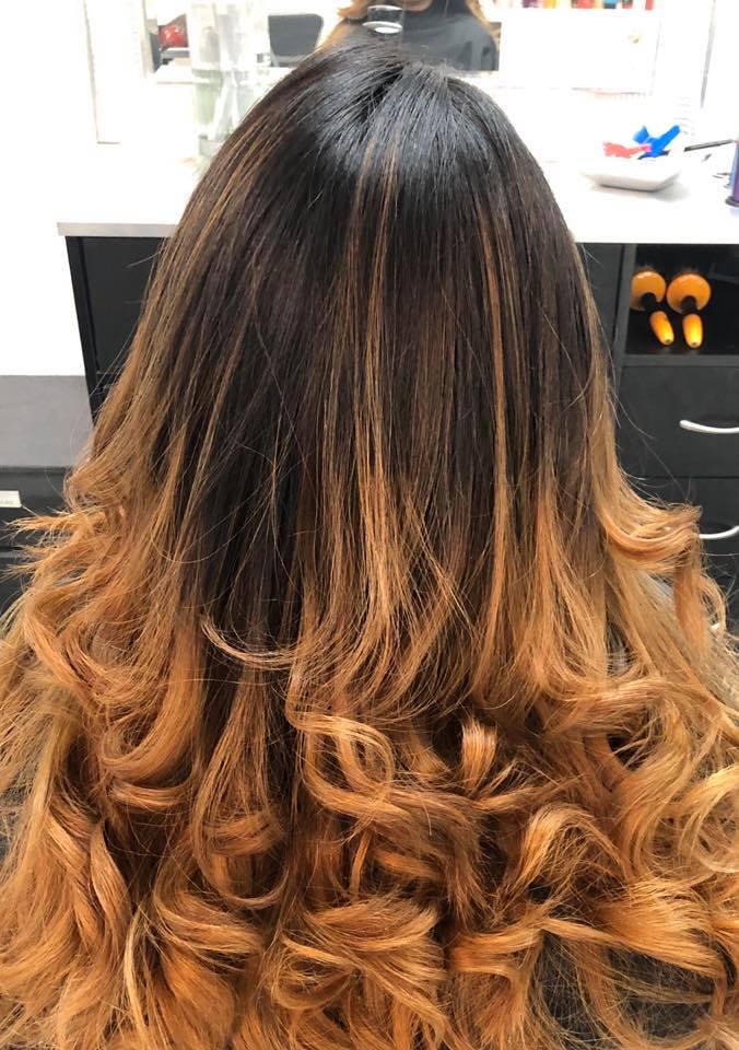 A woman with long curly hair is sitting in front of a mirror in a salon.