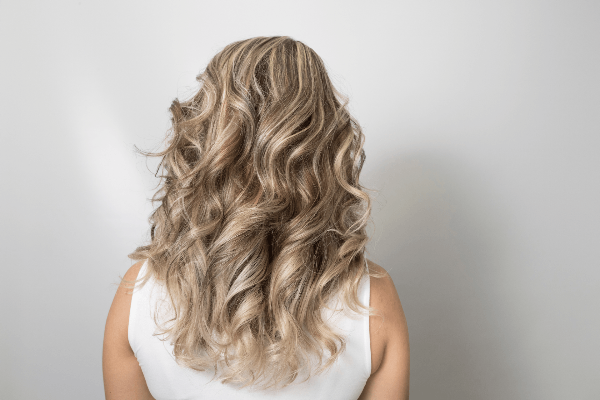 A woman with long blonde curly hair is standing in front of a white wall.