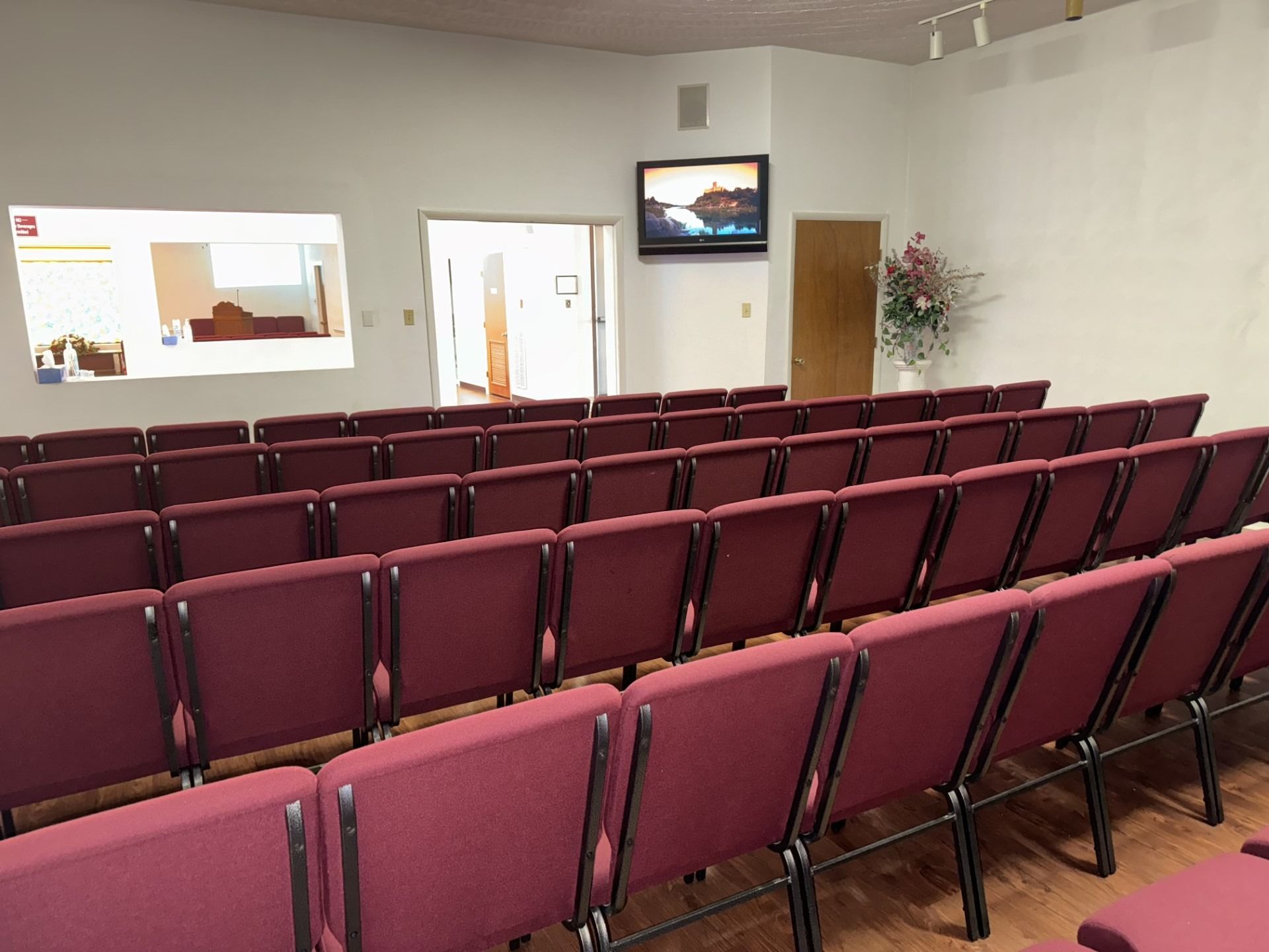 Rows of maroon chairs face a doorway and a television in a small, empty room.